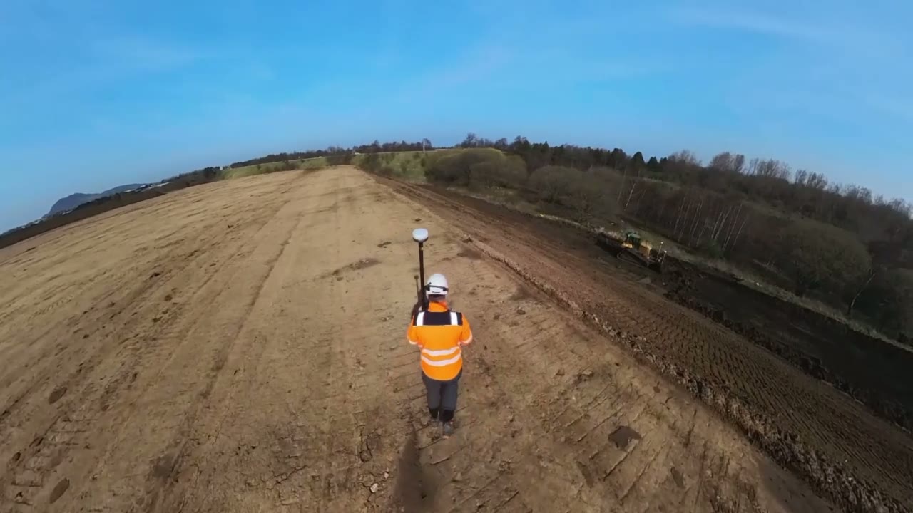 Stefan walking an earthworks platform with a GNSS rover pole while a dozer works alongside.