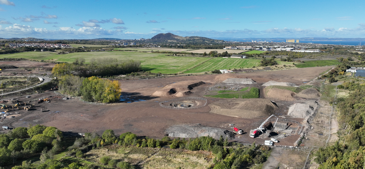 Aerial drone photo of the Shawfair Community Campus site during groundworks, with excavated areas, stockpiles and site cabins, Arthur's Seat on the horizon.