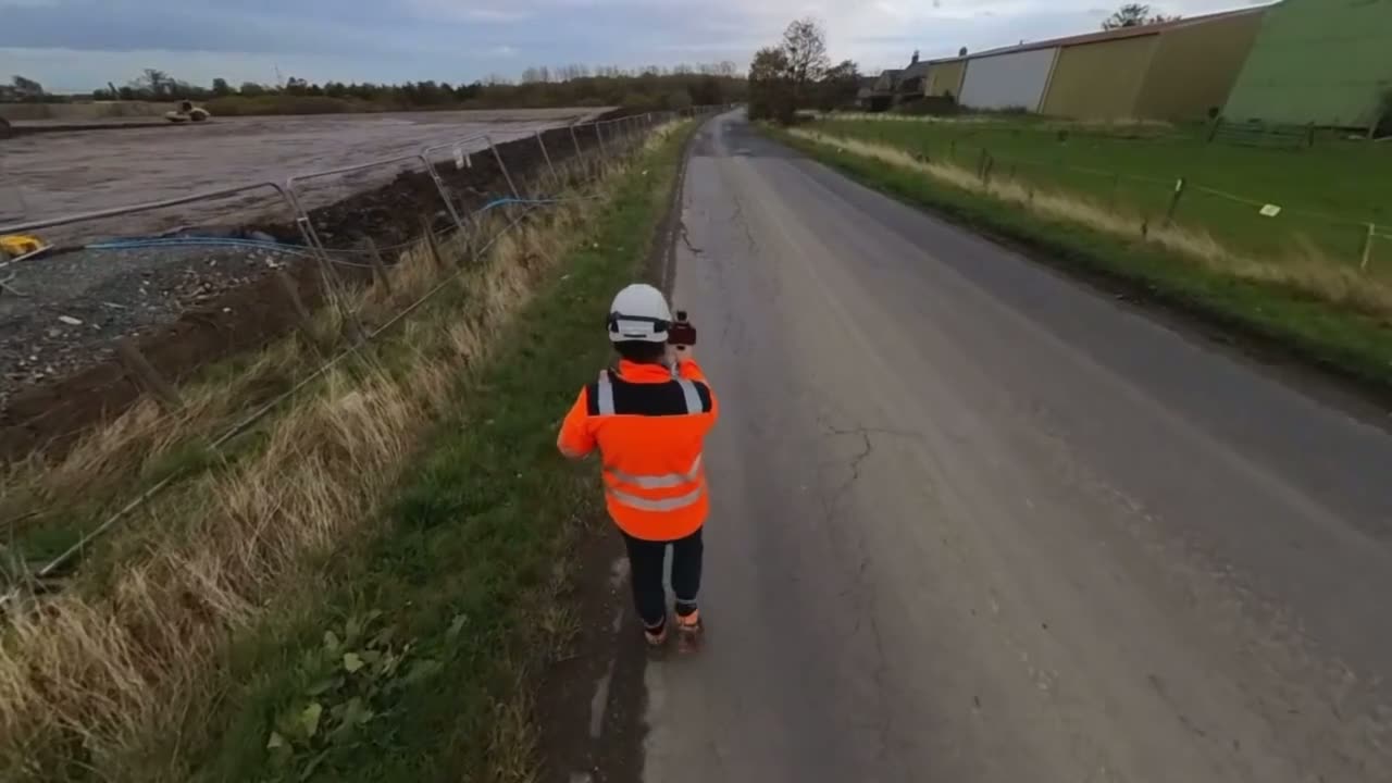 Stefan in hard hat and hi-vis walking a construction site boundary with a handheld SLAM laser scanner.